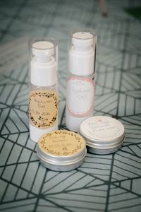 Handgemachte Kosmetikprodukte mit Etiketten &ndash; Ergebnisse eines Naturkosmetik-Workshops, einschlie&szlig;lich Hyaluron Gel und Anti-Age Balsam auf einem gemusterten Tisch.